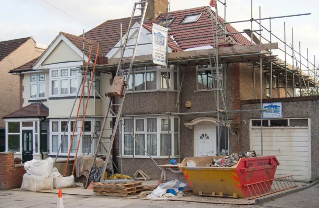Residential house under renovation with scaffolding, ladders, construction materials, and a skip on the driveway.