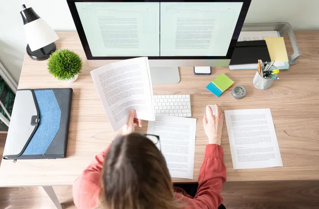 A woman at a desk sorting through papers while working on a computer, with stationery and office supplies nearby.