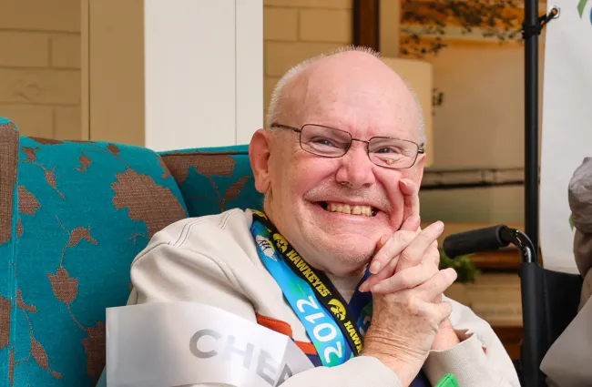 Joyful elderly man smiling while sitting in a vibrant chair, wearing a green tote bag and event lanyard.