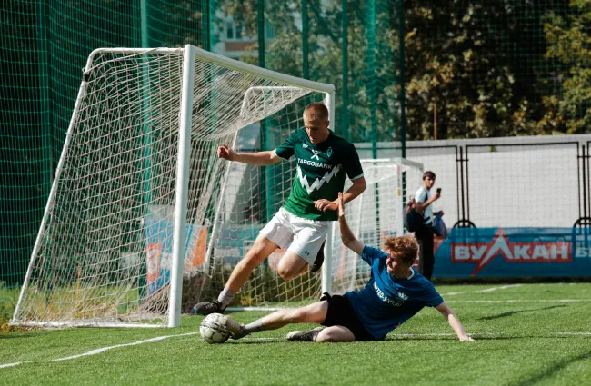 Two soccer players contesting a ball near the goalpost during a match on artificial turf.