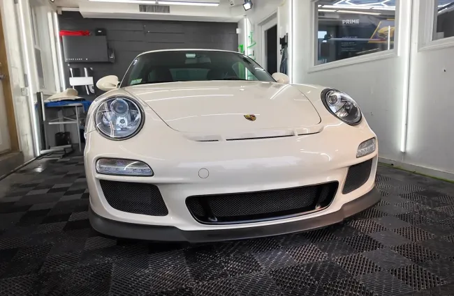 Front view of a white Porsche sports car parked indoors on a checkered floor with bright lighting.