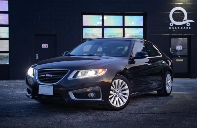 Black Saab sedan parked in front of a dark building with illuminated showroom windows at dusk.