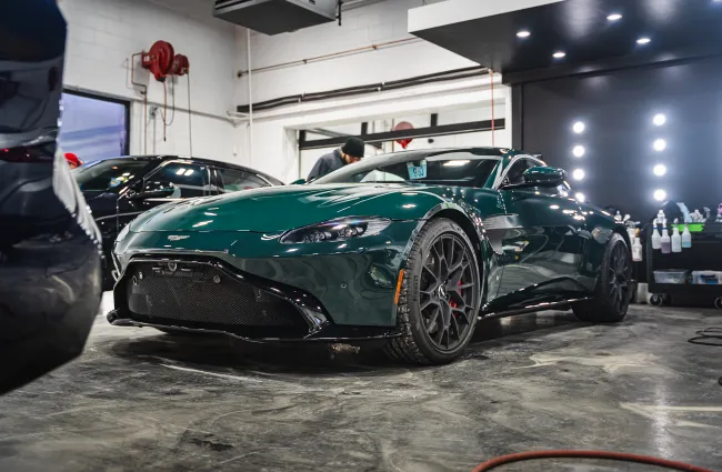 Dark green Aston Martin sports car in a brightly lit garage with polished concrete floor and cleaning supplies.