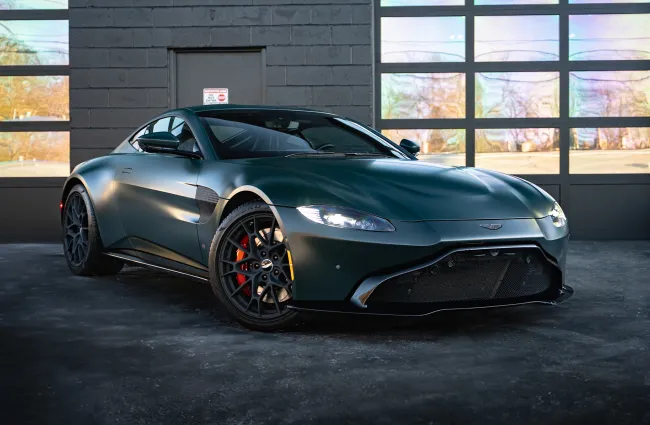 Matte dark green Aston Martin sports car parked indoors with black rims and red brake calipers.