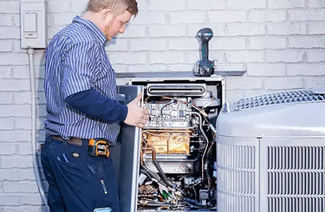 Technician inspecting and repairing an outdoor HVAC unit against a white brick wall background.