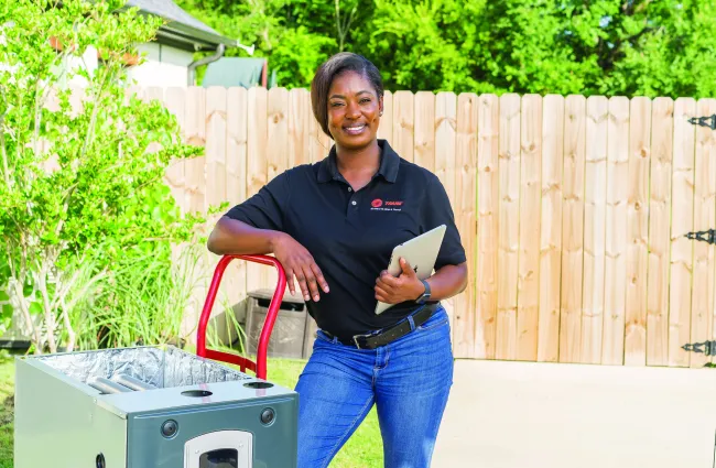 Technician in black shirt holding tablet smiles next to HVAC unit outdoors in bright sunny yard with wooden fence.