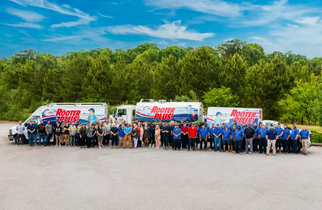 Large group of Rooter Plus plumbing team posing in front of service trucks with clear sky and green trees background.