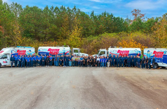 Large RooterPlus! plumbing team standing in front of five service trucks outdoors with trees in fall colors behind them.