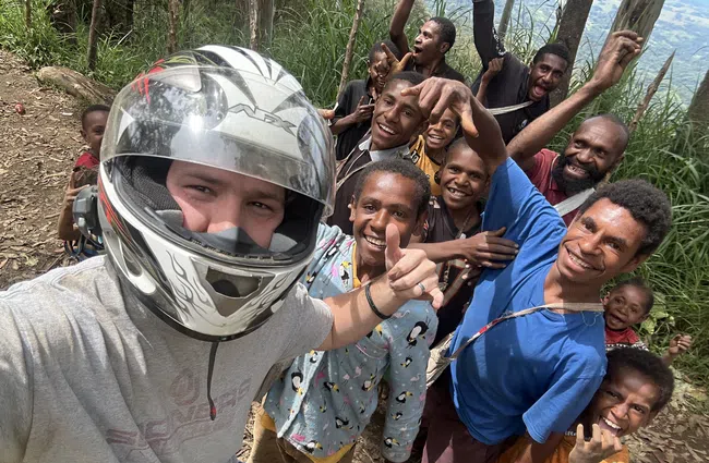 Group selfie of a person in a motorcycle helmet and local smiling people in a forested outdoor setting.