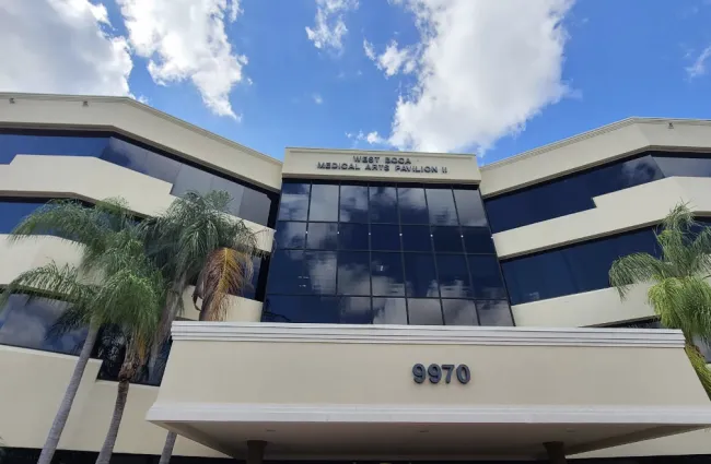 West Boca Medical Arts Pavilion II building with reflective windows and palm trees under a blue sky with clouds.