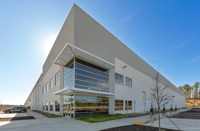 Modern industrial warehouse building with large glass entrance and surrounding parking lot under blue sky.