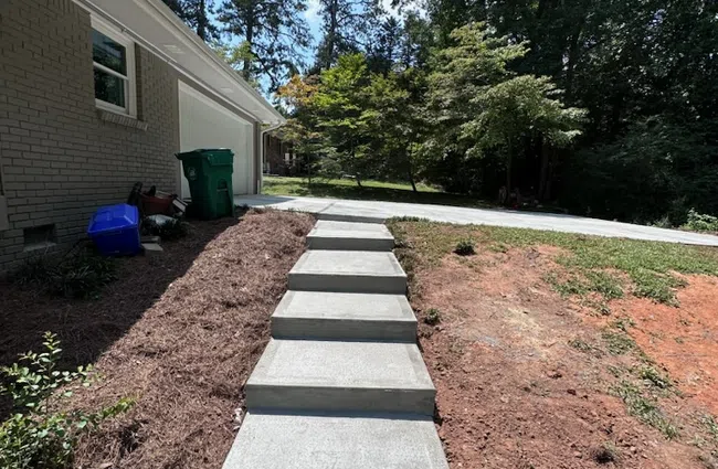 Curved driveway with cobblestone and concrete paving leading to a white colonial-style house surrounded by trees.