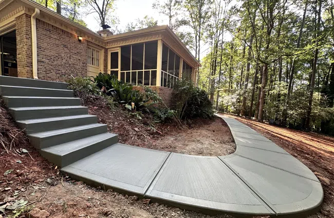 Newly poured concrete sidewalk and stairs leading up to a brick house surrounded by trees in a forested area.