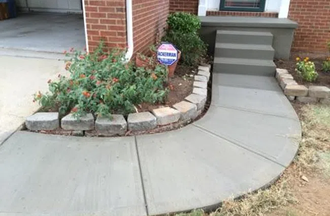 Curved concrete driveway with red brick-patterned section leading to a house surrounded by trees and landscaping.
