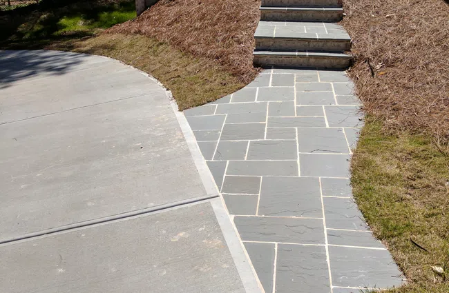 Stone walkway and steps leading from concrete driveway to elevated entrance with metal railing and landscaped yard.