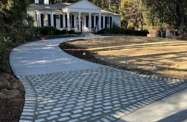 Curved driveway with cobblestone and concrete paving leading to a white colonial-style house surrounded by trees.