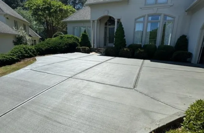 Freshly poured concrete driveway leading to a large white residential house with manicured shrubs and clear blue sky.
