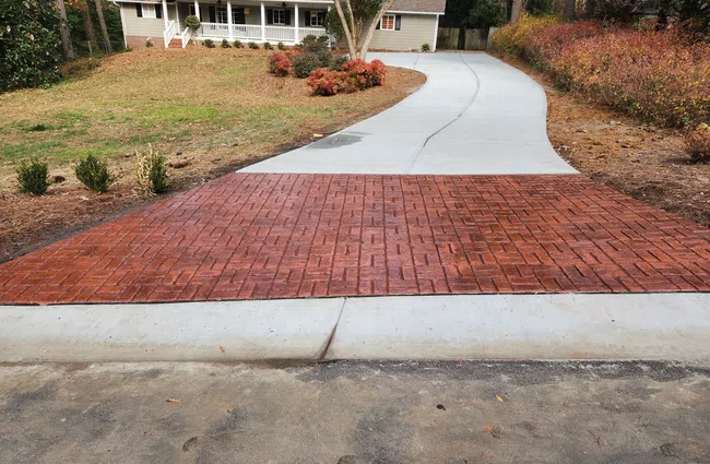 Curved concrete driveway with red brick-patterned section leading to a house surrounded by trees and landscaping.