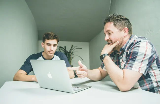 a couple of men sitting at a table looking at a laptop