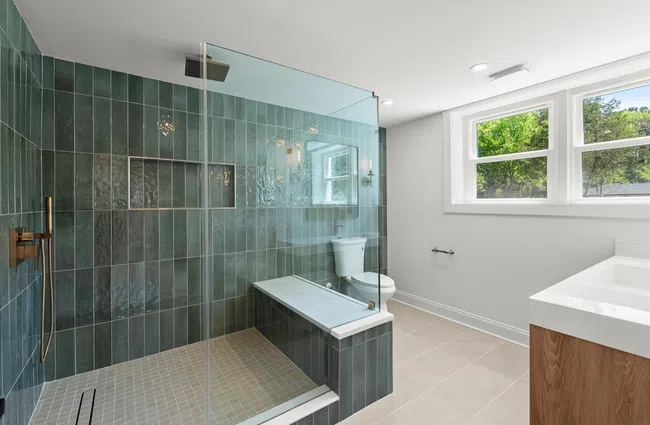 Modern bathroom with green tiled walk-in shower, built-in bench, white toilet, and large window with natural light.