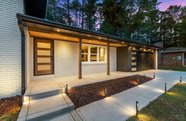 Modern house exterior with white brick, wooden door, garage, porch lights, and landscaped pathway at dusk.