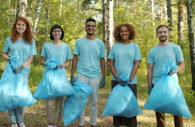 Group of five diverse volunteers in blue shirts holding blue trash bags during forest cleanup activity.