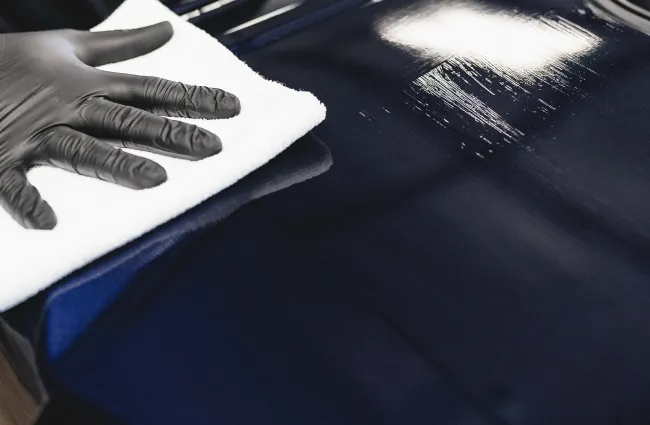 Person wearing black gloves polishing a shiny dark blue car surface with a white cloth indoors.