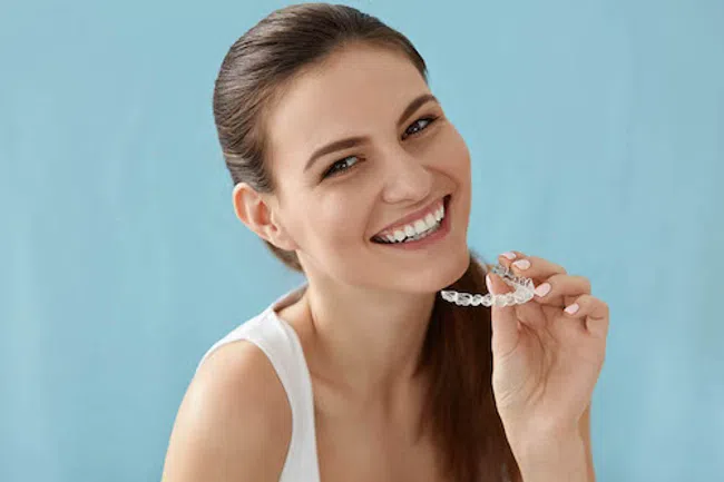 Smiling woman holding a clear dental aligner against a light blue background showing teeth care.