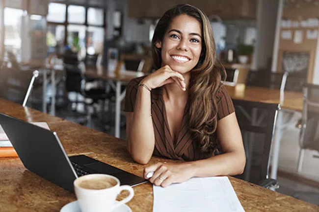 Smiling woman sitting by a laptop with coffee and documents in a modern office space.