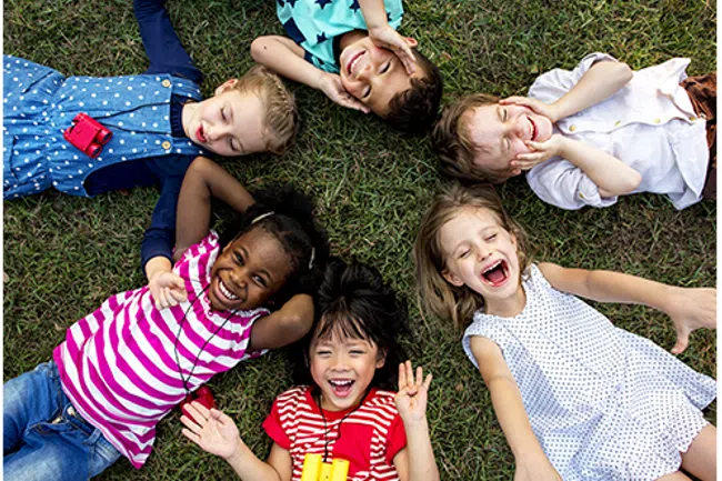 Six diverse children lying on grass in a circle, smiling and laughing on a sunny day.