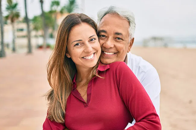 Smiling mature man hugging young woman in red sweater outdoors with palm trees and beach in background