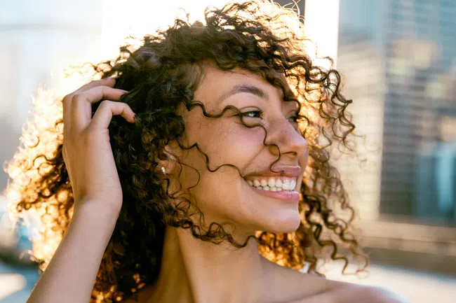 Smiling young woman with curly hair wearing a blue off-shoulder top in bright outdoor sunlight.