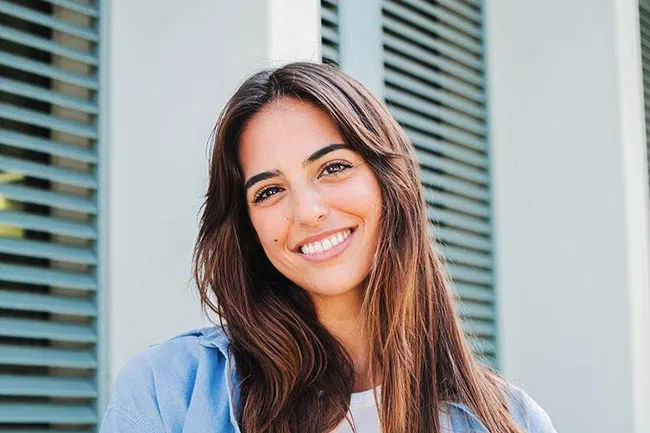 Smiling young woman with long hair wearing a blue shirt, standing in front of green shutters.