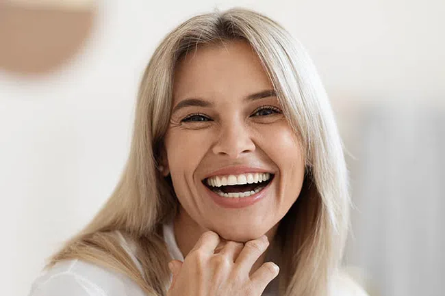 Smiling blonde woman with long hair wearing a white blouse, hand resting near chin, bright natural background