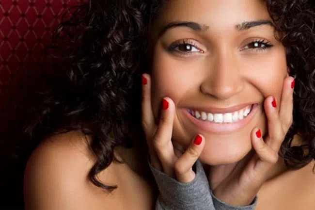 Close-up of a smiling woman with curly hair and red nail polish touching her face against a patterned background