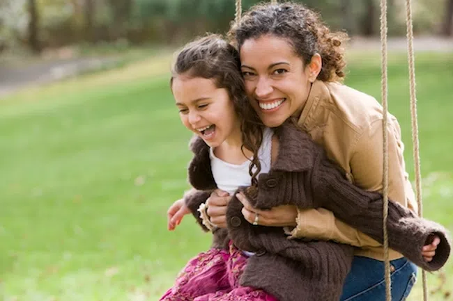 Mother and daughter smiling and enjoying time together on a swing in a green outdoor park setting.