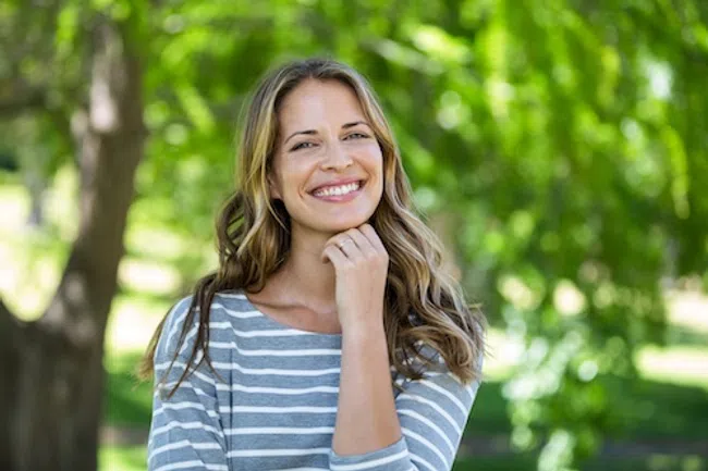 Smiling woman with long hair wearing a striped shirt outdoors with green trees in the background.