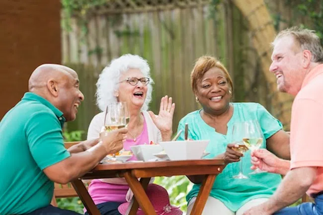 Four diverse seniors enjoying a meal and drinks together outdoors, laughing and socializing around a wooden table.