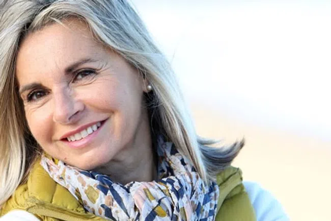 Smiling middle-aged woman with blonde hair wearing a patterned scarf and green jacket outdoors