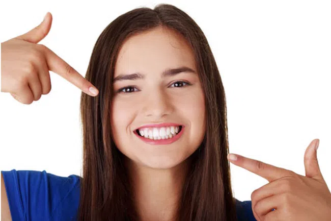Young woman with long brown hair smiling and pointing to her white teeth against white background