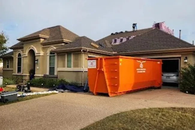 Residential house with roof under repair and large orange dumpster in driveway for construction debris removal.