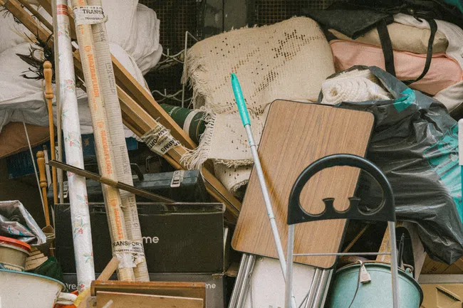 Pile of assorted household items and furniture stacked including a mop, boxes, folding table, and bags in a cluttered space