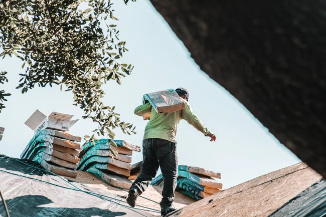 Worker carrying roofing materials on a sloped roof with stacked supplies under a blue sky and tree branches.