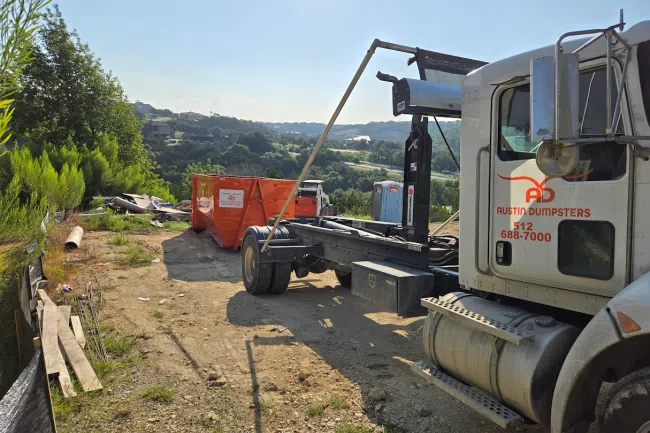 Austin Dumpsters truck unloading orange dumpster at a rural site with hills and greenery in the background.