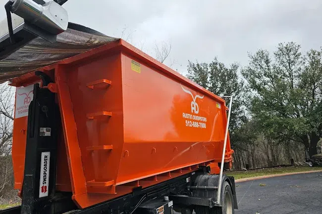 Large orange dumpster trailer with Austin Dumpsters branding parked on a driveway near trees and cloudy sky.