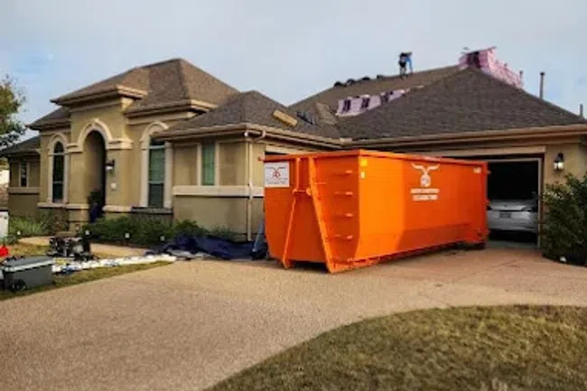 House with roof construction work in progress and large orange dumpster in driveway during daytime.