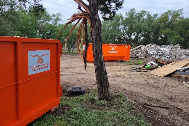 Two bright orange Austin Dumpsters at a debris cleanup site with trees and rubble in the background