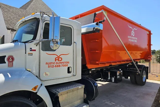 White and orange Austin Dumpsters truck parked outdoors under clear blue sky near a residential area.