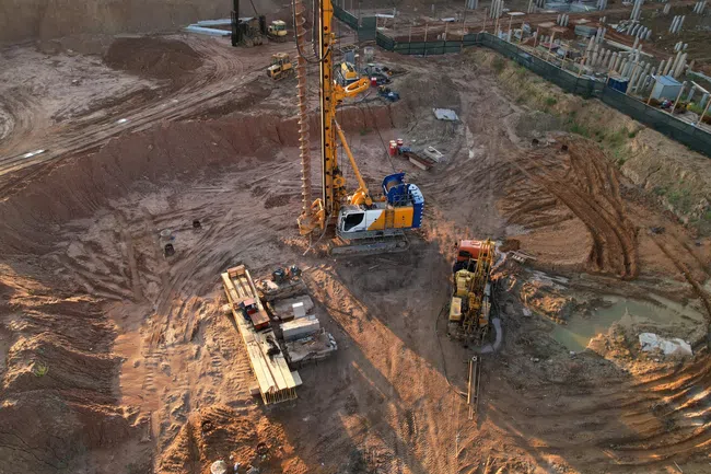 Aerial view of a construction site with heavy machinery drilling into the ground and building foundations nearby.