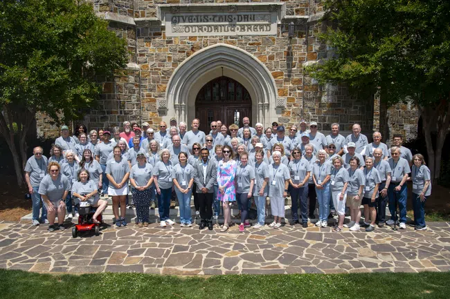 Large group of people in matching gray shirts posing outdoors in front of a stone building with an arched entrance.